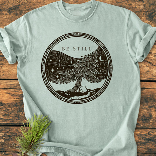1411 Be Still T-shirt: Pale green with a circular graphic of a pine tree, starry sky, and tent. BE STILL text above. Laid flat on wood, showcasing its relaxed fit and soft fabric.