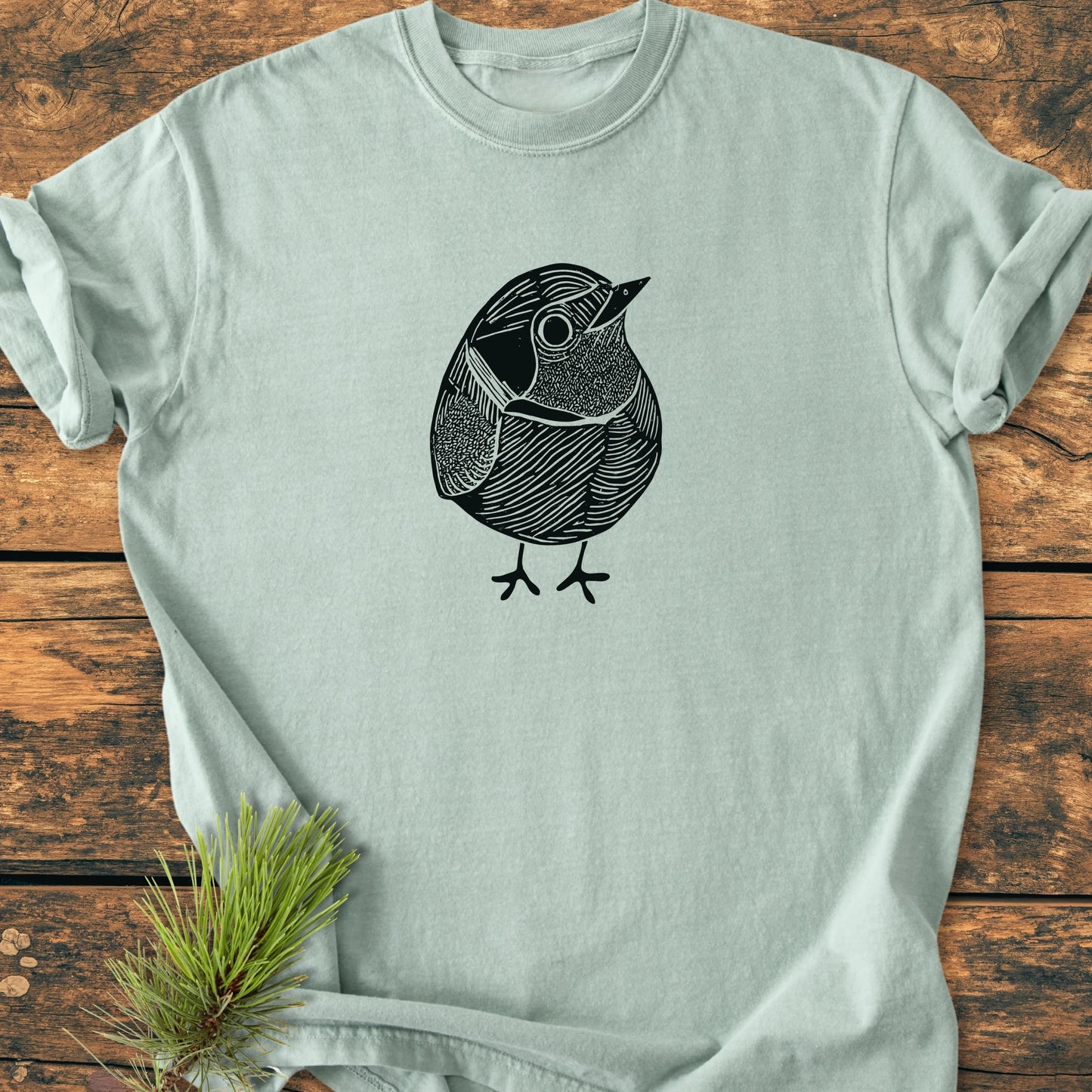 Light-sage crew-neck tee featuring a black line-art illustration of a small, round bird, titled 1433 Little Robin. Laid flat on a wooden surface with a pine needle sprig nearby.