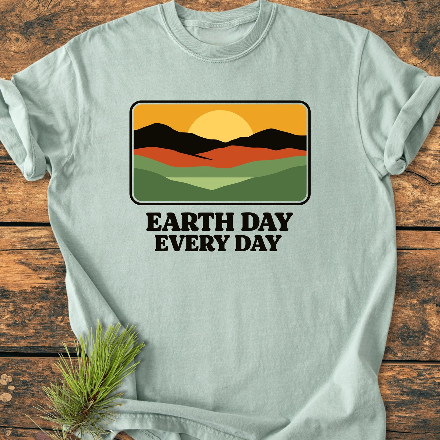 Pale green T-shirt with EARTH DAY EVERY DAY text and a sunrise landscape graphic, emphasizing eco-consciousness. Laid on wood with a pine sprig, highlighting its nature-inspired design.