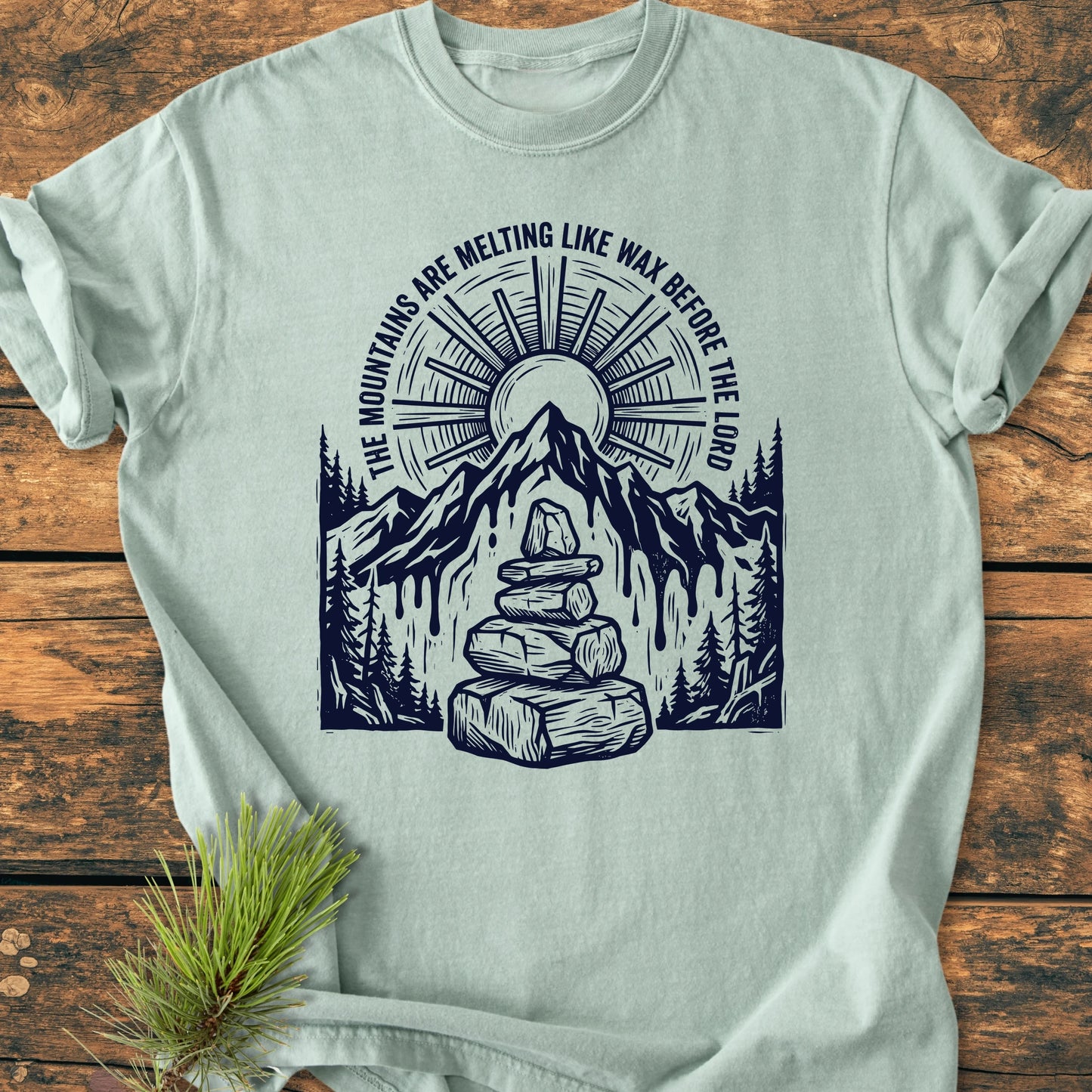 Pale T-shirt with 1414 Mountains Melting Like Wax design, featuring a sun, mountain, pine trees, and cairn. Woodcut-style, relaxed fit, next to a pine sprig on wooden surface.