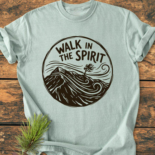 Folded 1410 Walk In The Spirit T-shirt featuring a circular dark-brown graphic with a mountain landscape and tree, styled with a pine sprig on a wooden surface.