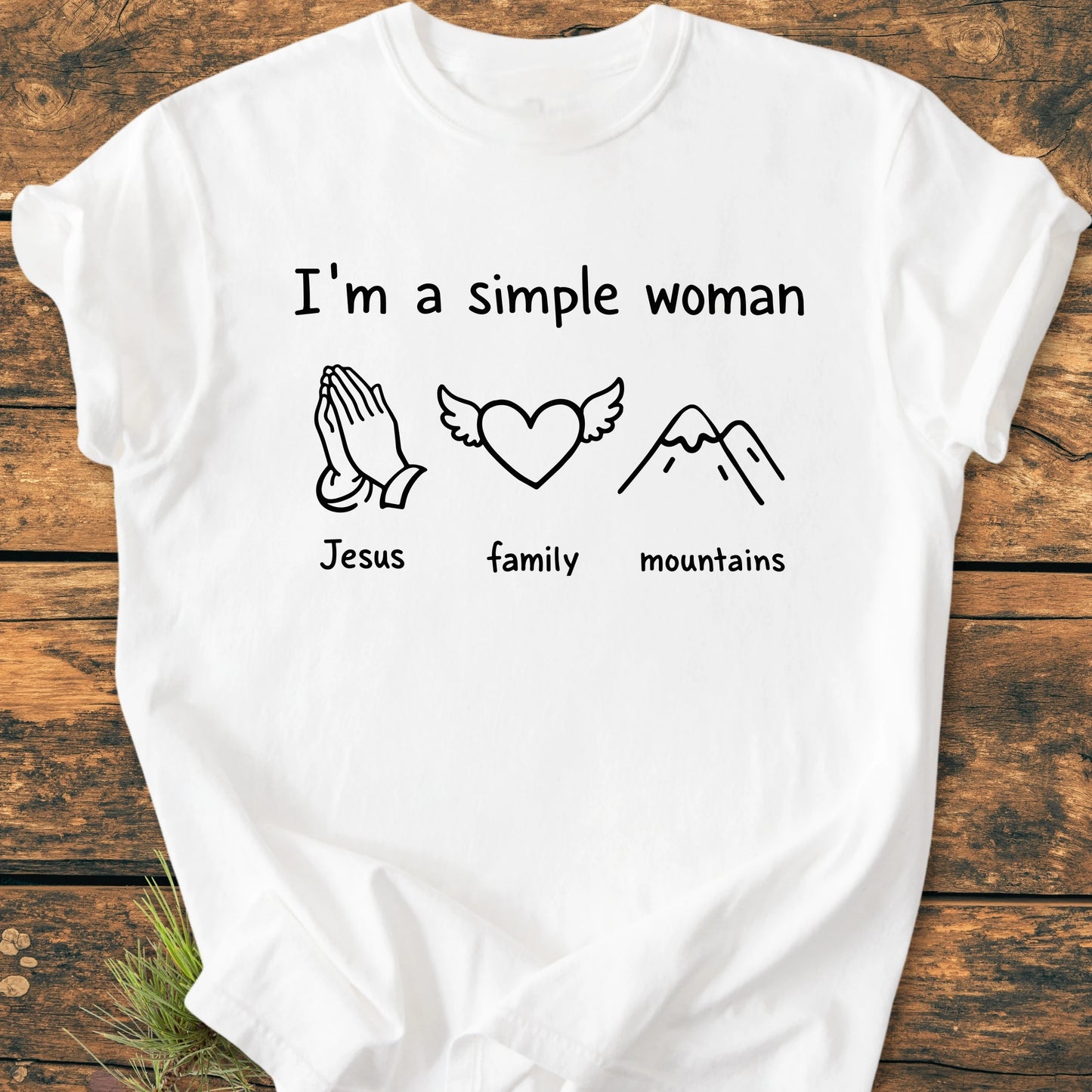 White T-shirt with I'm a simple woman in black font, featuring line-drawings: praying hands labeled Jesus, heart with wings labeled family, and stylized mountains labeled mountains, on a wooden surface.