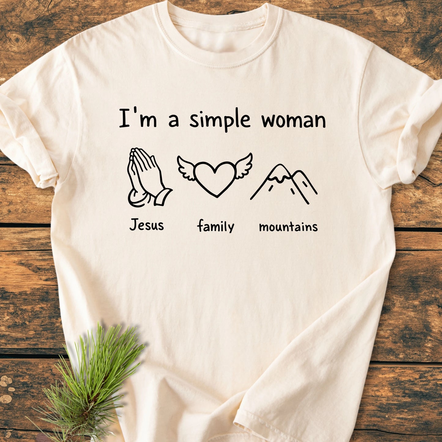 Light-colored T-shirt with I'm a Simple Woman above icons of praying hands, heart with wings, and mountains, on a wooden surface with pine sprig, showcasing its vintage wash texture.
