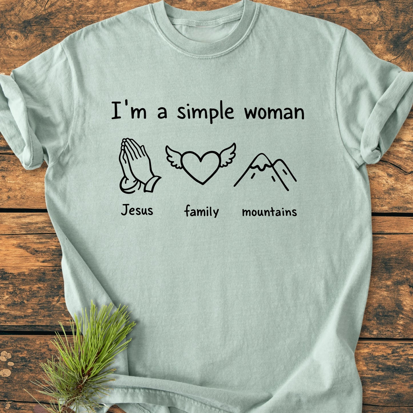 Pale-blue T-shirt with I'm a Simple Woman text, featuring icons: praying hands labeled Jesus, heart with wings labeled family, mountain outline labeled mountains. Laid flat on a wooden surface.